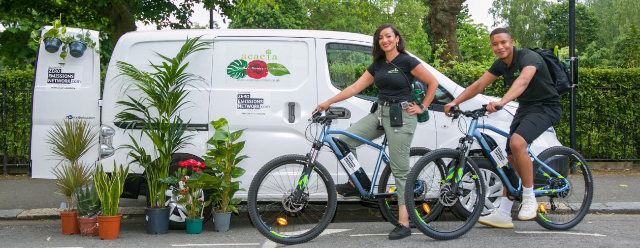 A woman and a man stand next to bikes, a van and plants