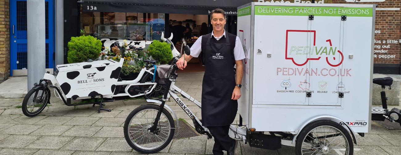 A smiling man stands in front of a large cargo bike which says 'Pedivan' on it