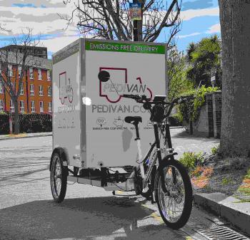 A large white cargo trike - with a big box on the back and the word 'Pedivan' on it on the road