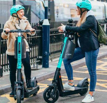 Two women wearing blue helmets pose on TIER e-scooters